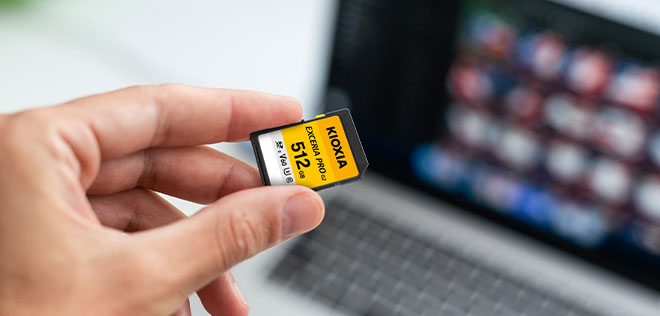 A hand holding the EXCERIA PRO G2 SD Card in front of a laptop